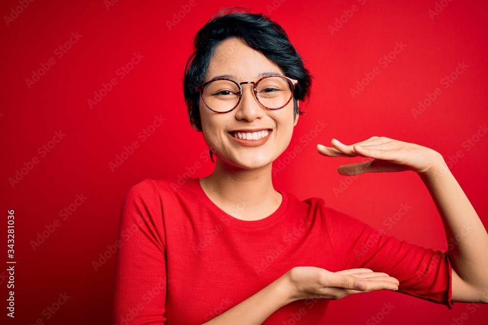 Young beautiful asian girl wearing casual t-shirt and glasses over isolated red background gesturing with hands showing big and large size sign, measure symbol. Smiling looking at the camera. Measure Young beautiful asian girl wearing casual t-shirt and glasses over isolated red background gesturing with hands showing big and large size sign, measure symbol. Smiling looking at the camera. Measure