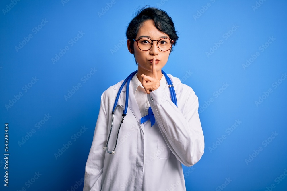Young beautiful asian doctor girl wearing stethoscope and coat with blue cancer ribbon asking to be quiet with finger on lips. Silence and secret concept. Young beautiful asian doctor girl wearing stethoscope and coat with blue cancer ribbon asking to be quiet with finger on lips. Silence and secret concept.