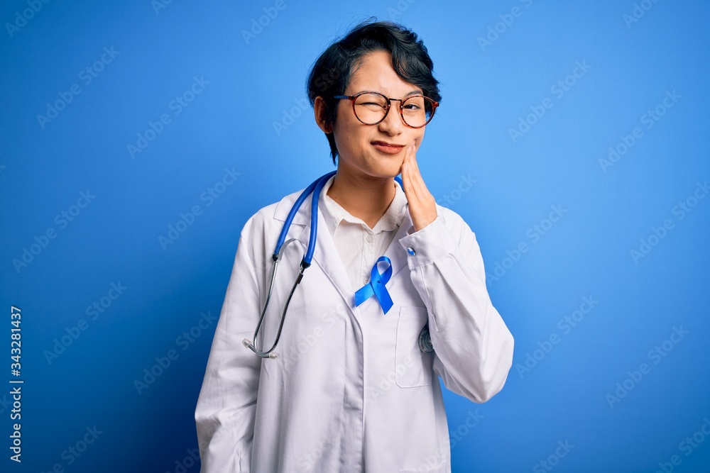 Young beautiful asian doctor girl wearing stethoscope and coat with blue cancer ribbon touching mouth with hand with painful expression because of toothache or dental illness on teeth. Dentist Young beautiful asian doctor girl wearing stethoscope and coat with blue cancer ribbon touching mouth with hand with painful expression because of toothache or dental illness on teeth. Dentist