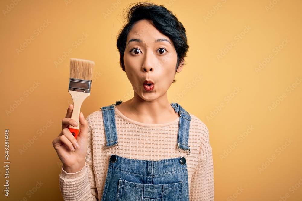 Young beautiful chinese woman painting holding paint brush over isolated yellow background scared in shock with a surprise face, afraid and excited with fear expression Young beautiful chinese woman painting holding paint brush over isolated yellow background scared in shock with a surprise face, afraid and excited with fear expression