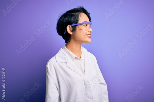 Murais de parede Young beautiful asian scientist girl wearing coat and glasses over purple background looking away to side with smile on face, natural expression