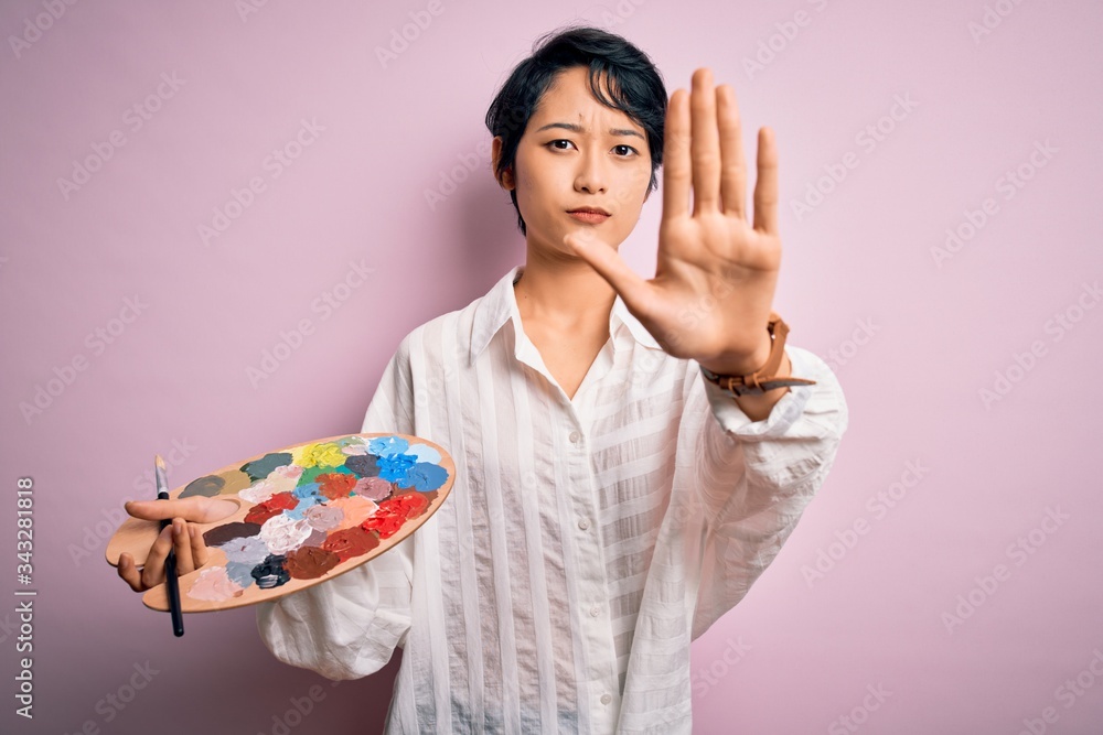 Young beautiful chinese artist woman painting using paintbrush and palette with colors with open hand doing stop sign with serious and confident expression, defense gesture Young beautiful chinese artist woman painting using paintbrush and palette with colors with open hand doing stop sign with serious and confident expression, defense gesture