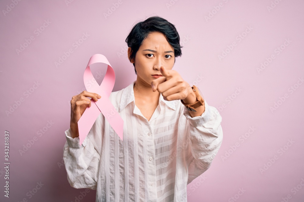 Young beautiful asian girl holding pink cancer ribbon symbol over isolated background pointing with finger to the camera and to you, hand sign, positive and confident gesture from the front Young beautiful asian girl holding pink cancer ribbon symbol over isolated background pointing with finger to the camera and to you, hand sign, positive and confident gesture from the front