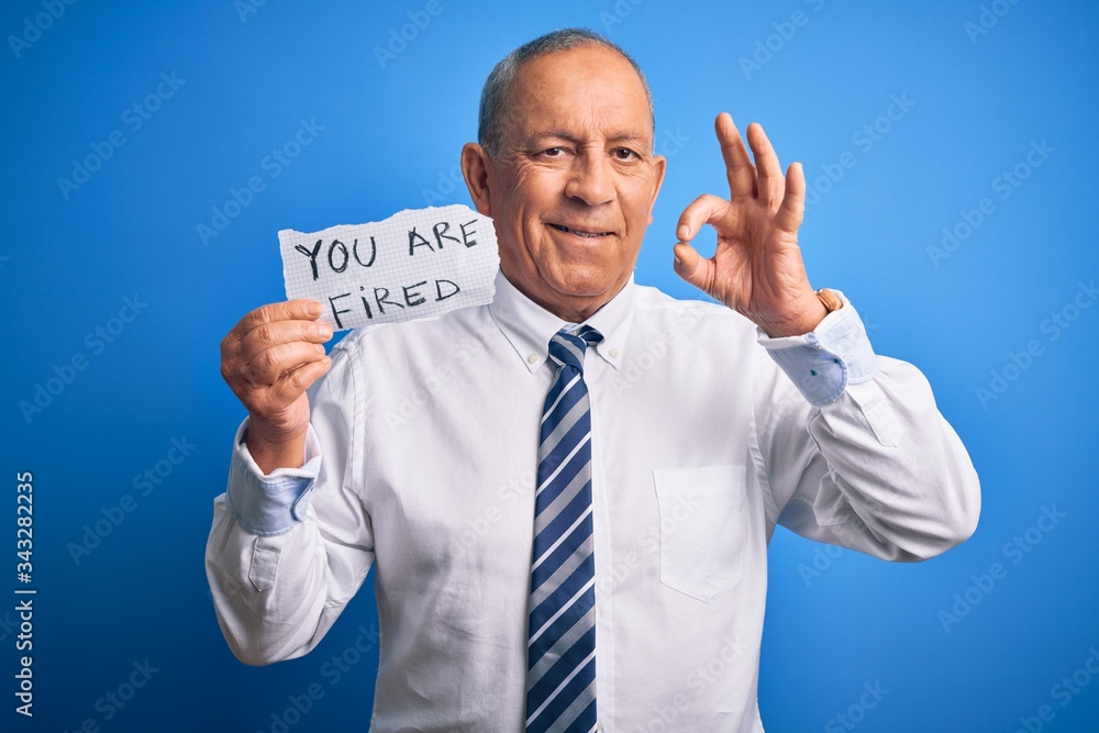 Senior handsome businessman holding you are fired paper over isolated blue background doing ok sign with fingers, excellent symbol Senior handsome businessman holding you are fired paper over isolated blue background doing ok sign with fingers, excellent symbol