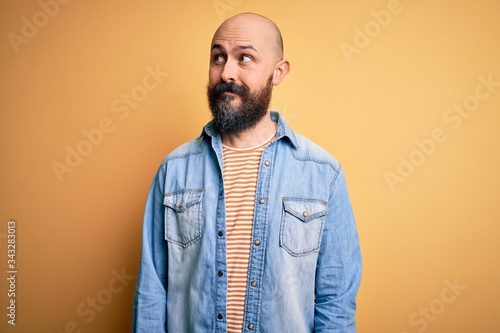 Handsome bald man with beard wearing casual denim jacket and striped t-shirt smiling looking to the side and staring away thinking.