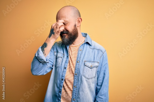 Handsome bald man with beard wearing casual denim jacket and striped t-shirt tired rubbing nose and eyes feeling fatigue and headache. Stress and frustration concept.