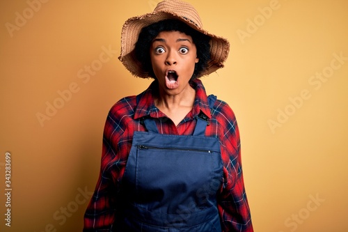 Young African American afro farmer woman with curly hair wearing apron and hat afraid and shocked with surprise expression, fear and excited face.