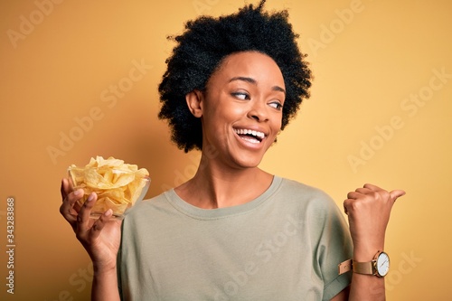 Young African American afro woman with curly hair holding bowl with chips potatoes pointing and showing with thumb up to the side with happy face smiling