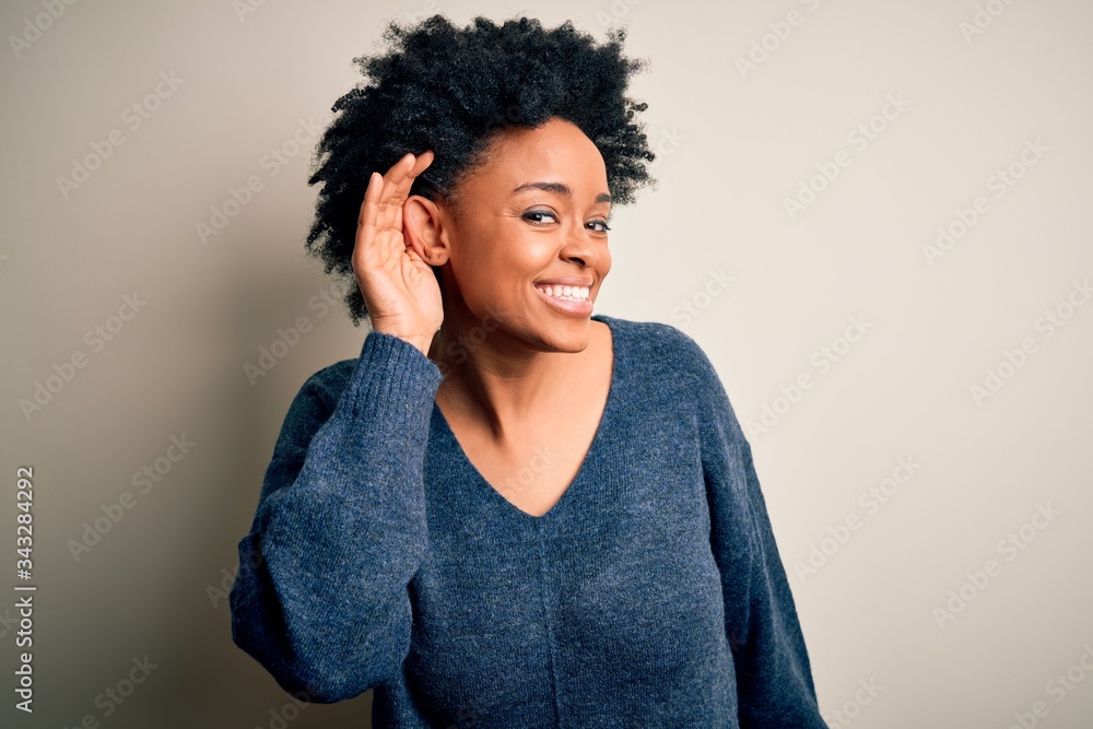 Young beautiful African American afro woman with curly hair wearing casual sweater smiling with hand over ear listening an hearing to rumor or gossip. Deafness concept. Young beautiful African American afro woman with curly hair wearing casual sweater smiling with hand over ear listening an hearing to rumor or gossip. Deafness concept.