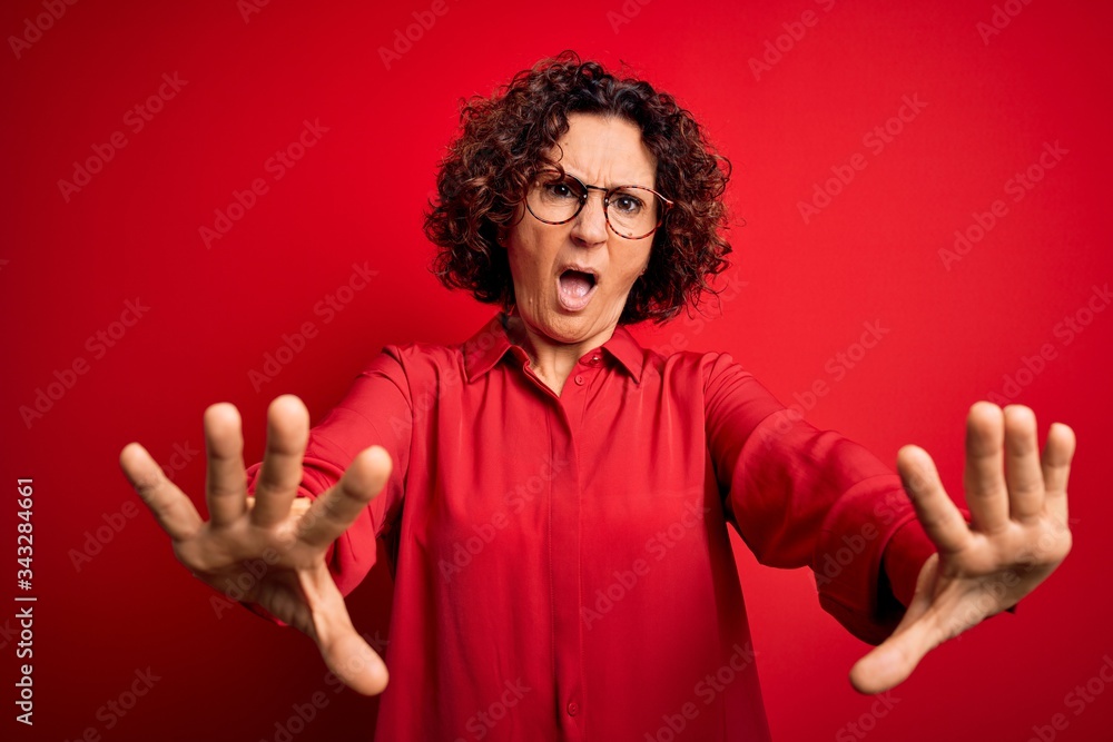 Middle age beautiful curly hair woman wearing casual shirt and glasses over red background doing stop gesture with hands palms, angry and frustration expression Middle age beautiful curly hair woman wearing casual shirt and glasses over red background doing stop gesture with hands palms, angry and frustration expression
