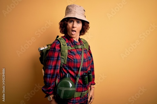 Middle age curly hair hiker woman hiking wearing backpack and water canteen using binoculars In shock face, looking skeptical and sarcastic, surprised with open mouth