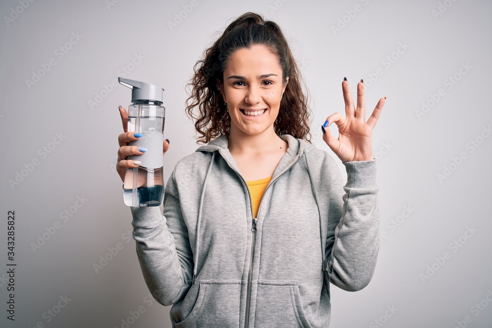 Beautiful woman with curly hair doing sport drinking bottle of water over white background doing ok sign with fingers, excellent symbol Beautiful woman with curly hair doing sport drinking bottle of water over white background doing ok sign with fingers, excellent symbol