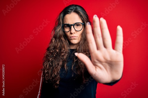 Young beautiful woman with curly hair wearing sweater and glasses over red background doing stop sing with palm of the hand. Warning expression with negative and serious gesture on the face.
