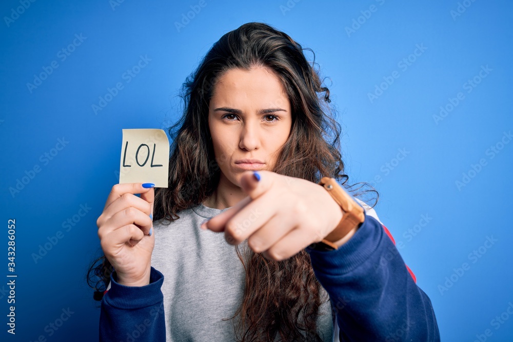 Naklejka premium Young beautiful woman with curly hair holding reminder paper with lol message pointing with finger to the camera and to you, hand sign, positive and confident gesture from the front