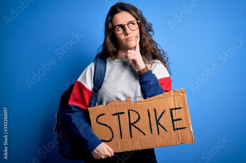 Beautiful student woman with curly hair wearing backpack holding banner with strike message serious face thinking about question, very confused idea © Krakenimages.com