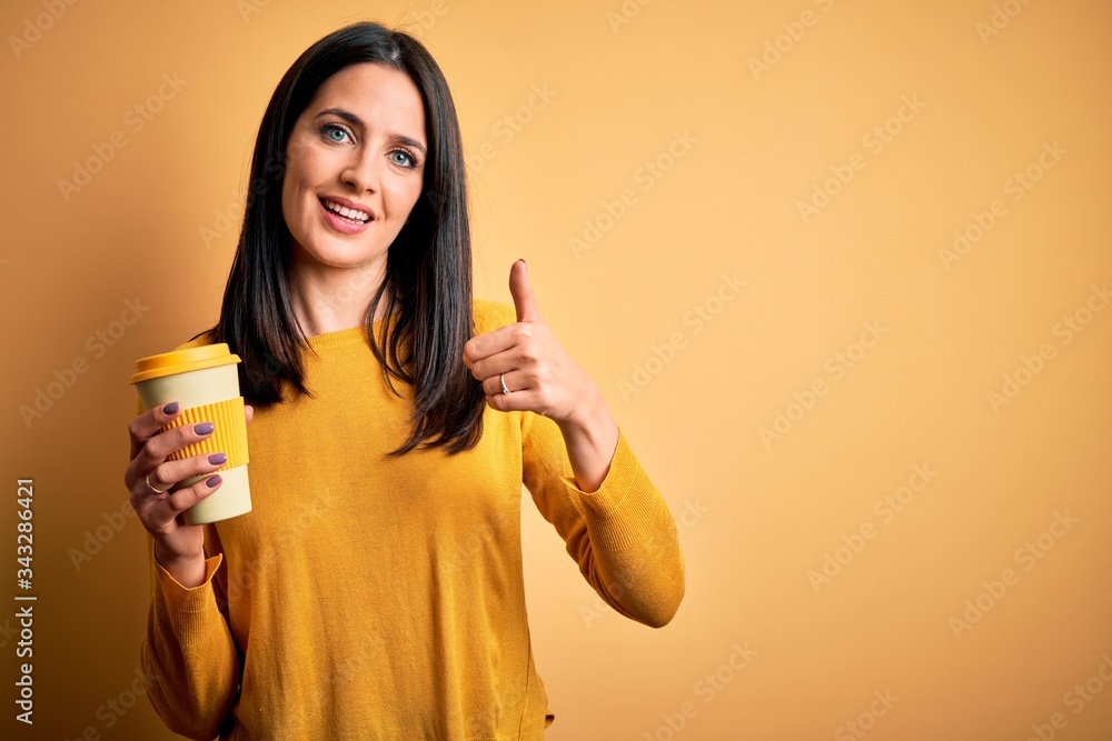 Young woman with blue eyes holding cup of coffee standing over yellow background happy with big smile doing ok sign, thumb up with fingers, excellent sign Young woman with blue eyes holding cup of coffee standing over yellow background happy with big smile doing ok sign, thumb up with fingers, excellent sign