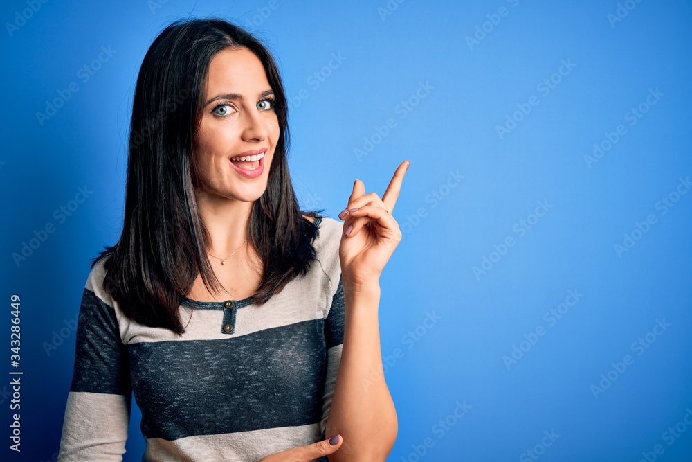 Young brunette woman with blue eyes wearing casual striped t-shirt standing over background with a big smile on face, pointing with hand and finger to the side looking at the camera. Young brunette woman with blue eyes wearing casual striped t-shirt standing over background with a big smile on face, pointing with hand and finger to the side looking at the camera.