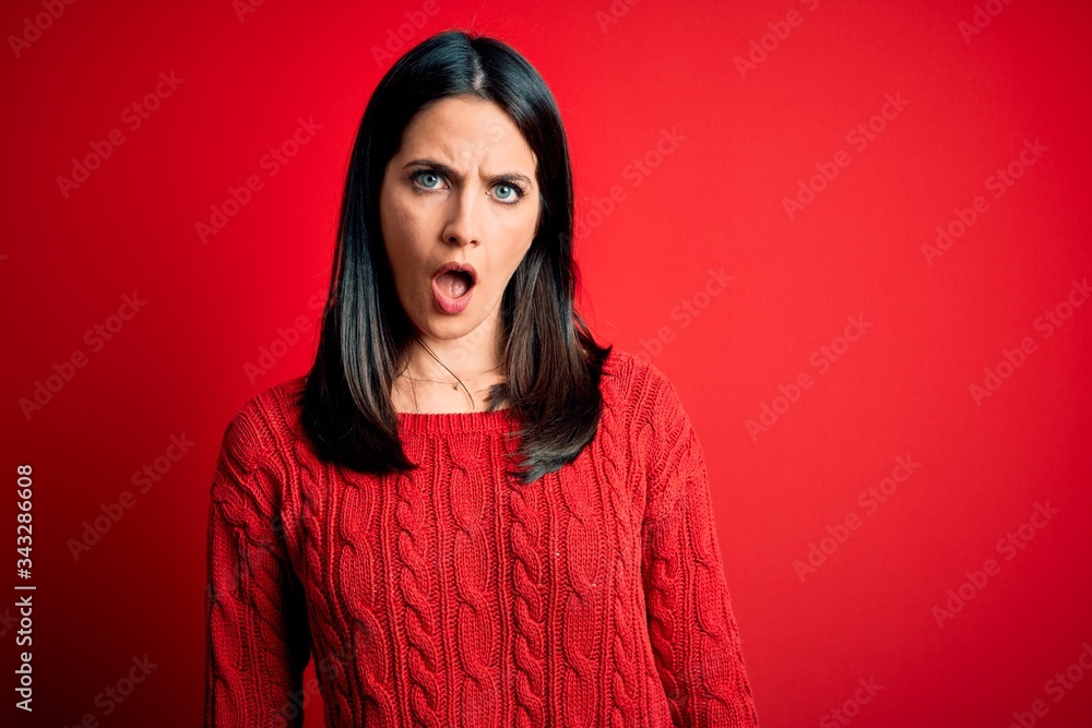 Fototapeta premium Young brunette woman with blue eyes wearing casual sweater over isolated red background In shock face, looking skeptical and sarcastic, surprised with open mouth