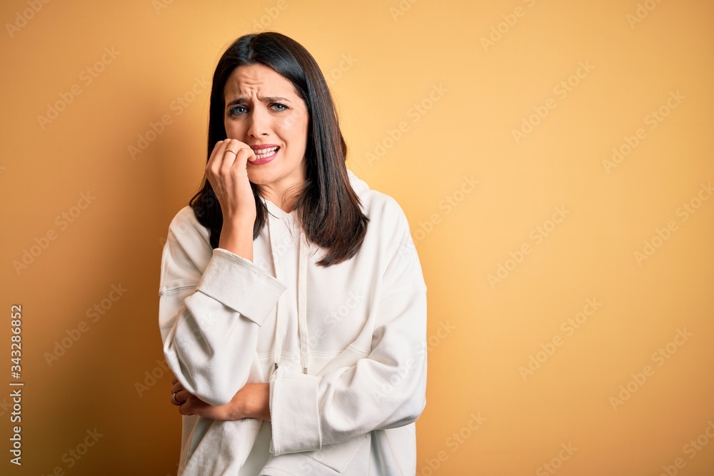 Young brunette sportswoman with blue eyes wearing training sweatshirt over yellow background looking stressed and nervous with hands on mouth biting nails. Anxiety problem.