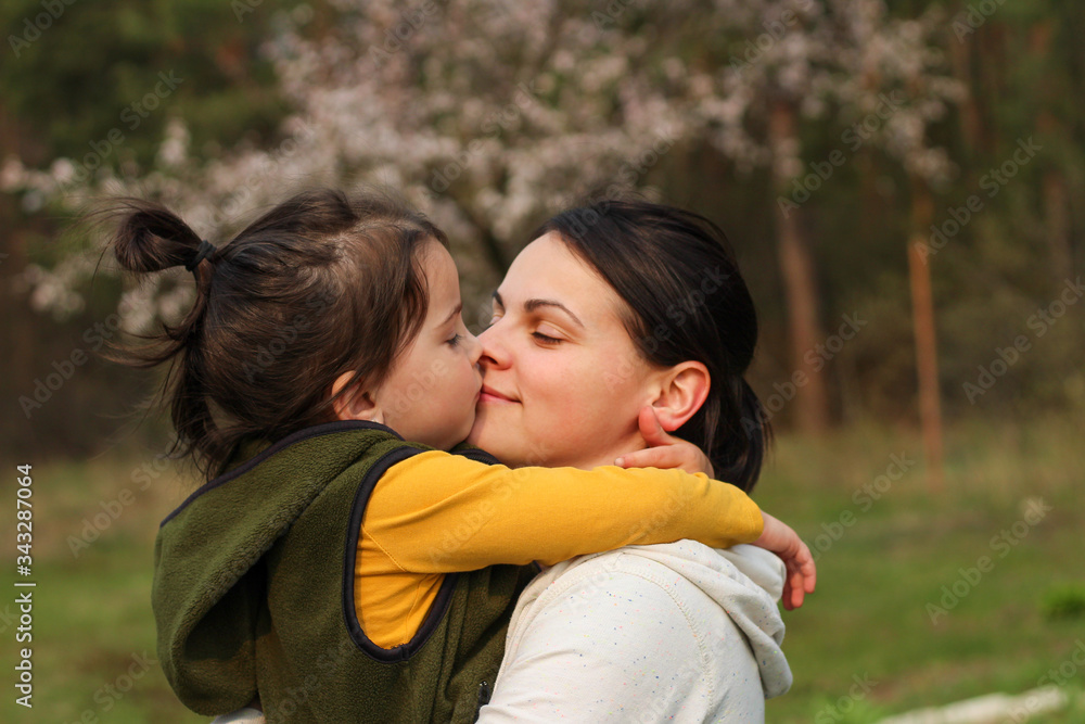 Beautiful mother and daughter hugging and kissing in casual clothes. Warm baby hug on the background of a blurry flowering tree Beautiful mother and daughter hugging and kissing in casual clothes. Warm baby hug on the background of a blurry flowering tree