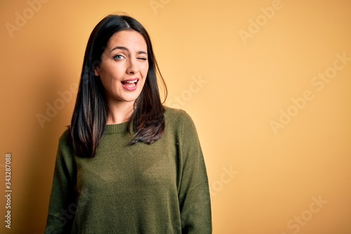 Young brunette woman with blue eyes wearing green casual sweater over yellow background winking looking at the camera with sexy expression, cheerful and happy face. © Krakenimages.com