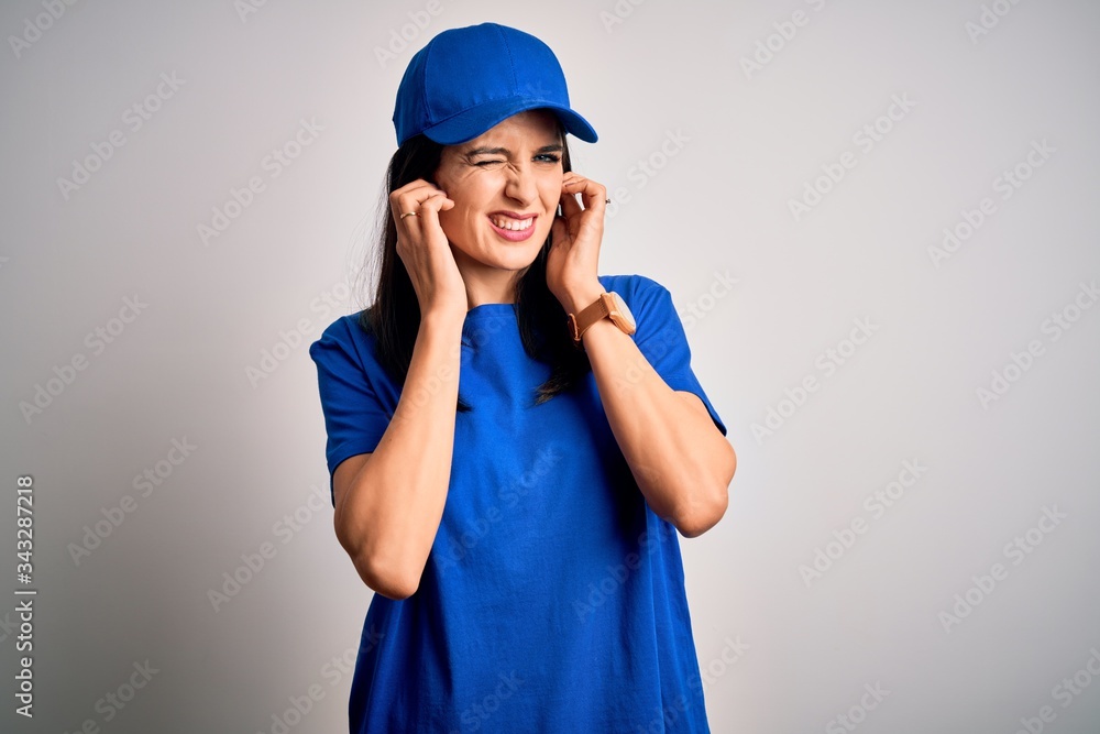 Young delivery woman with blue eyes wearing cap standing over blue background covering ears with fingers with annoyed expression for the noise of loud music. Deaf concept. Young delivery woman with blue eyes wearing cap standing over blue background covering ears with fingers with annoyed expression for the noise of loud music. Deaf concept.