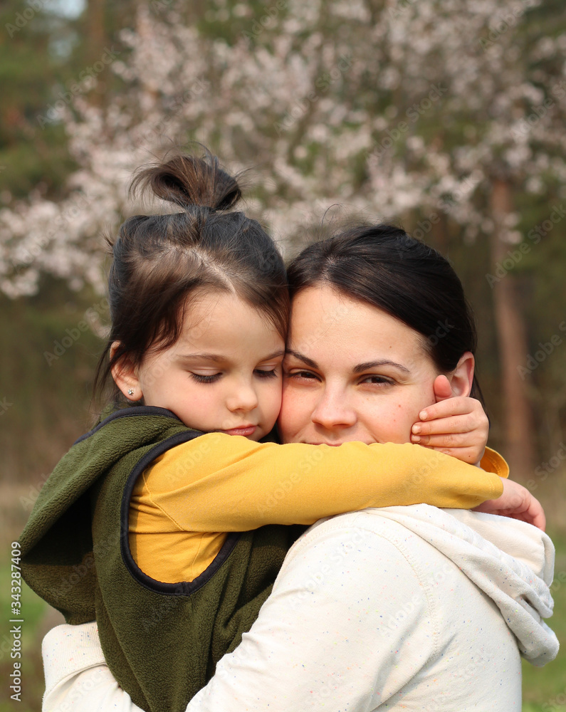 Beautiful mother and daughter hugging and kissing in casual clothes. Warm baby hug on the background of a blurry flowering tree Beautiful mother and daughter hugging and kissing in casual clothes. Warm baby hug on the background of a blurry flowering tree