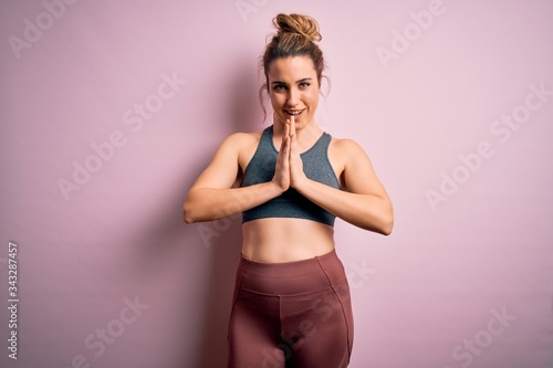 Young beautiful blonde sportswoman doing sport wearing sportswear over pink background praying with hands together asking for forgiveness smiling confident.