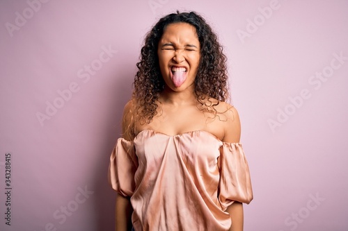 Young beautiful woman with curly hair wearing casual t-shirt standing over pink background sticking tongue out happy with funny expression. Emotion concept.