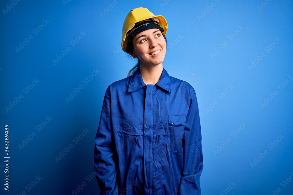 Young beautiful worker woman with blue eyes wearing security helmet and uniform looking away to side with smile on face, natural expression. Laughing confident. Young beautiful worker woman with blue eyes wearing security helmet and uniform looking away to side with smile on face, natural expression. Laughing confident.