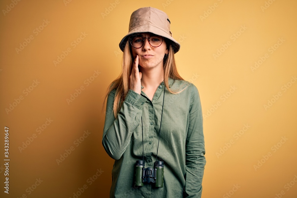 Beautiful blonde explorer woman with blue eyes wearing hat and glasses using binoculars touching mouth with hand with painful expression because of toothache or dental illness on teeth. Dentist