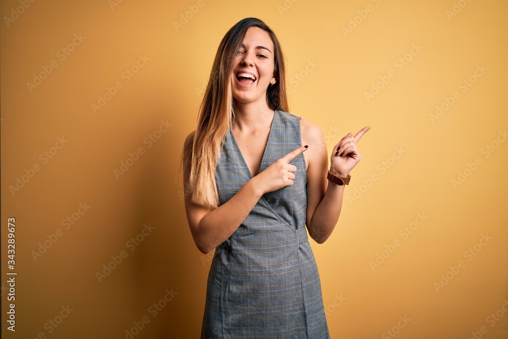 Young beautiful blonde woman with blue eyes wearing dress over yellow background smiling and looking at the camera pointing with two hands and fingers to the side. Young beautiful blonde woman with blue eyes wearing dress over yellow background smiling and looking at the camera pointing with two hands and fingers to the side.