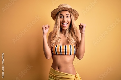Young beautiful blonde woman on vacation wearing bikini and hat over yellow background celebrating surprised and amazed for success with arms raised and open eyes. Winner concept.
