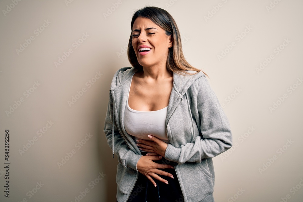 Young beautiful brunette sportswoman wearing sportswoman training over white background with hand on stomach because nausea, painful disease feeling unwell. Ache concept. Young beautiful brunette sportswoman wearing sportswoman training over white background with hand on stomach because nausea, painful disease feeling unwell. Ache concept.