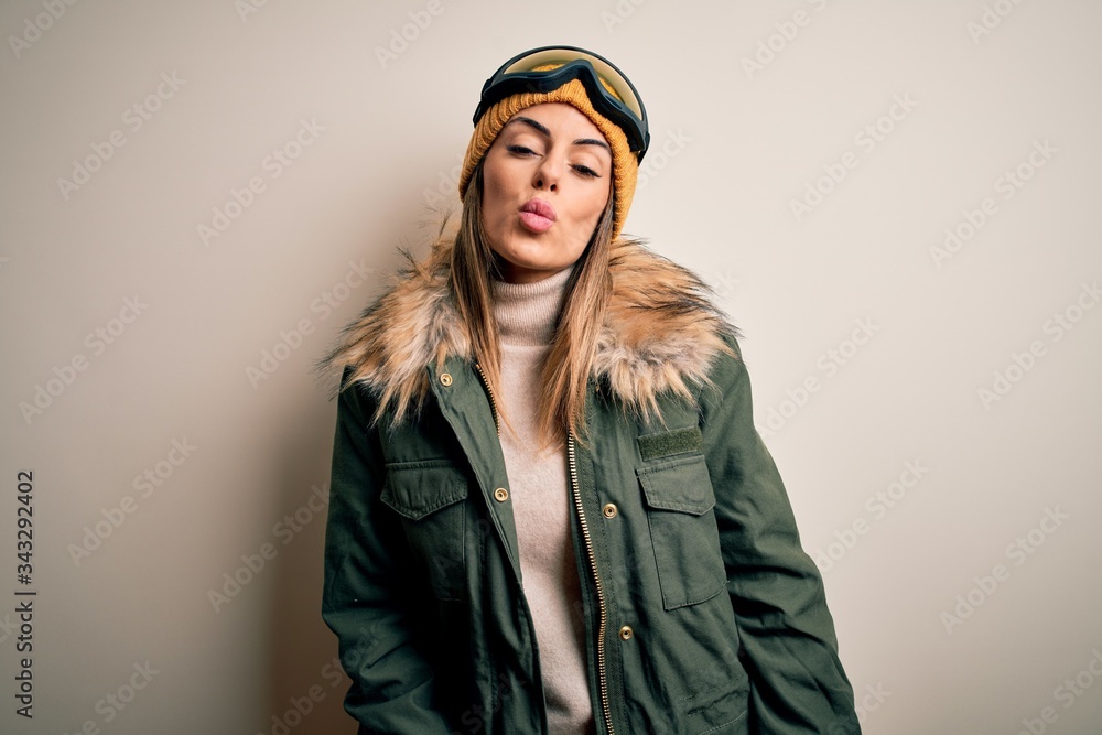 Young brunette skier woman wearing snow clothes and ski goggles over white background looking at the camera blowing a kiss on air being lovely and sexy. Love expression.