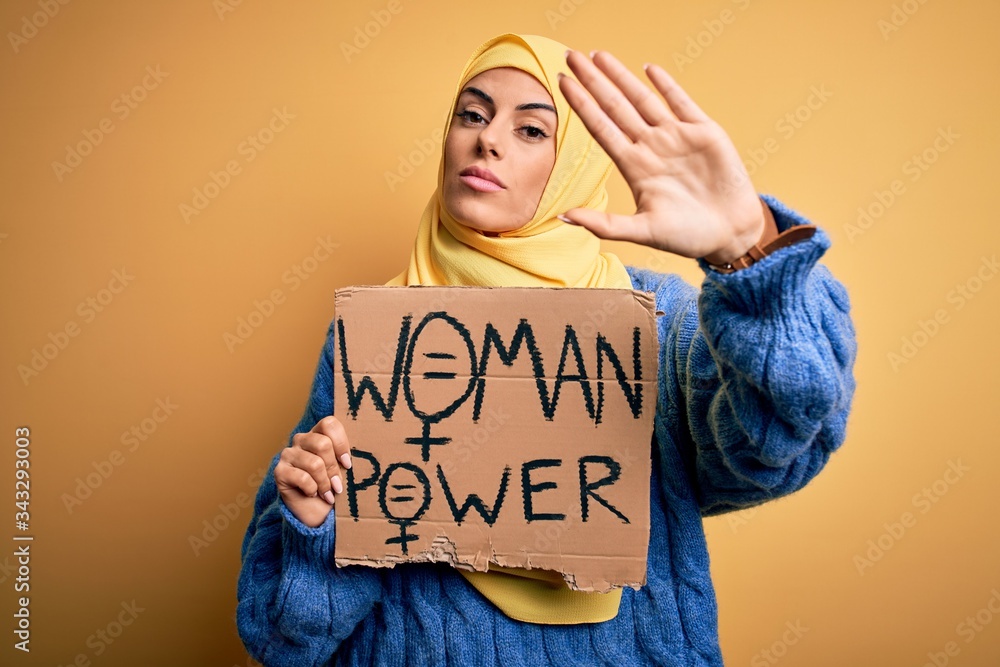 Young beautiful arab woman wearing islamic hijab holding banner with women power message with open hand doing stop sign with serious and confident expression, defense gesture Young beautiful arab woman wearing islamic hijab holding banner with women power message with open hand doing stop sign with serious and confident expression, defense gesture