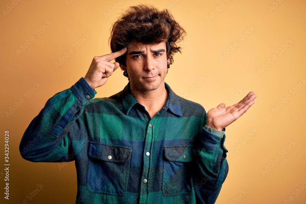 Young handsome man wearing casual shirt standing over isolated yellow background confused and annoyed with open palm showing copy space and pointing finger to forehead. Think about it. Young handsome man wearing casual shirt standing over isolated yellow background confused and annoyed with open palm showing copy space and pointing finger to forehead. Think about it.