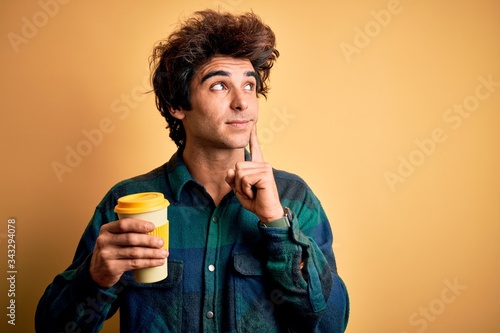 Young handsome man drinking glass of coffee standing over isolated yellow background serious face thinking about question, very confused idea © Krakenimages.com