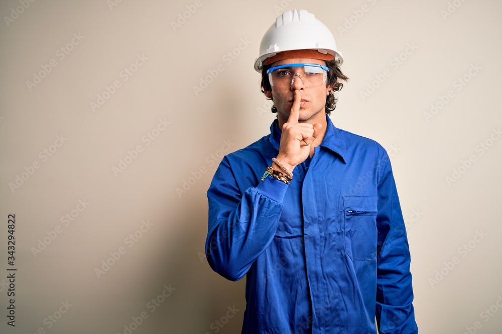 Young constructor man wearing uniform and security helmet over isolated white background asking to be quiet with finger on lips. Silence and secret concept.
