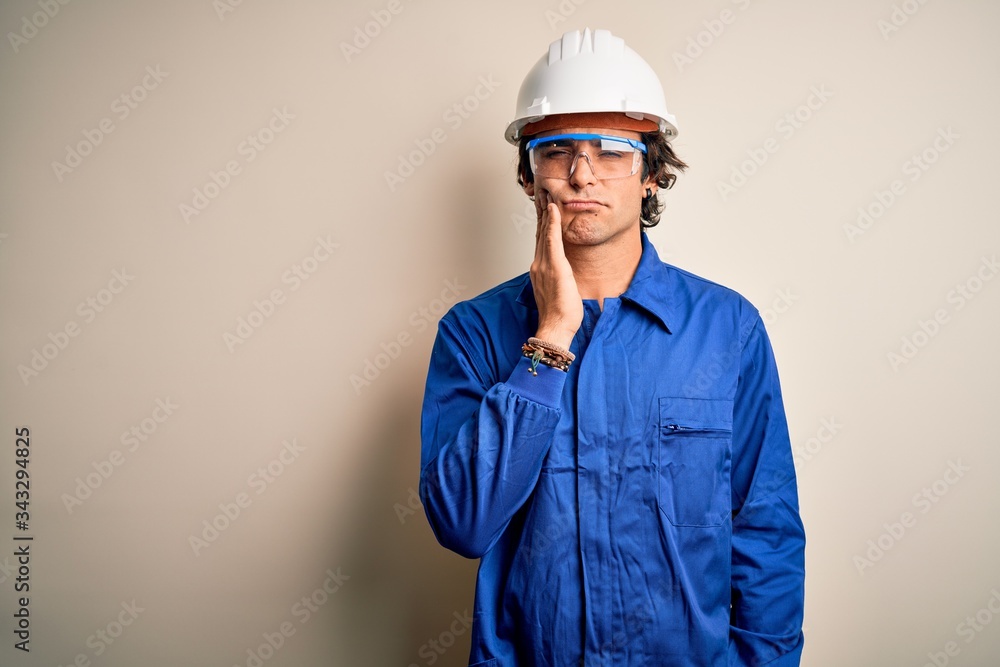 Young constructor man wearing uniform and security helmet over isolated white background touching mouth with hand with painful expression because of toothache or dental illness on teeth. Dentist
