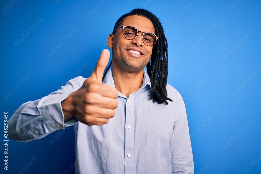 Young handsome african american man with dreadlocks wearing casual shirt and glasses doing happy thumbs up gesture with hand. Approving expression looking at the camera showing success. Young handsome african american man with dreadlocks wearing casual shirt and glasses doing happy thumbs up gesture with hand. Approving expression looking at the camera showing success.
