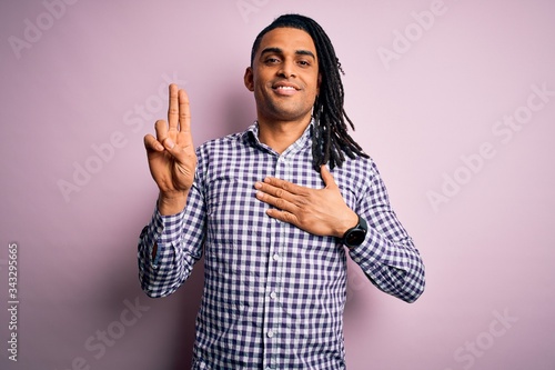 Young handsome african american afro man with dreadlocks wearing casual shirt smiling swearing with hand on chest and fingers up, making a loyalty promise oath