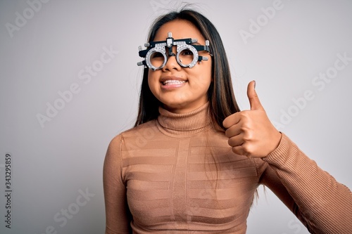 Young asian optical girl controlling eyesight using optometry glasses over white background doing happy thumbs up gesture with hand. Approving expression looking at the camera showing success.