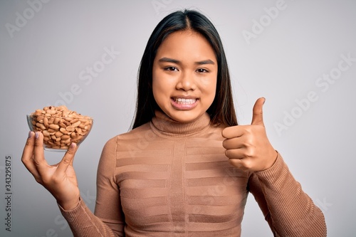 Young asian girl holding bowl of healthy peanuts eating as snack for diet over white background happy with big smile doing ok sign, thumb up with fingers, excellent sign