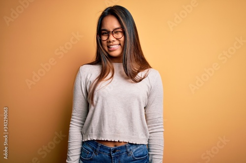 Young beautiful asian girl wearing casual sweater and glasses over yellow background winking looking at the camera with sexy expression, cheerful and happy face.