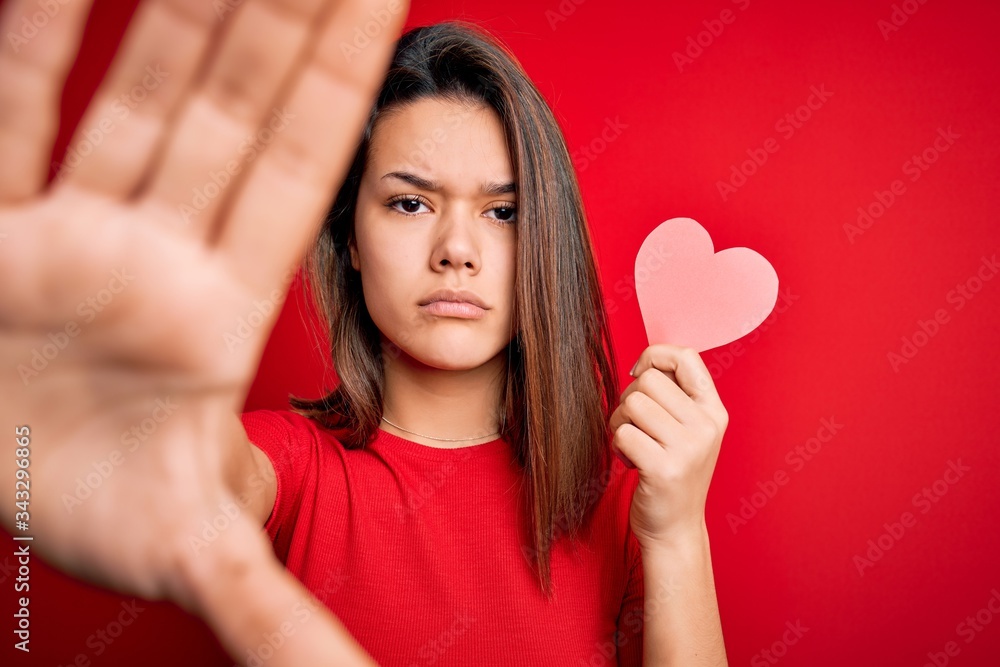 Young beautiful brunette romantic girl holding red paper heart shape over isolated background with open hand doing stop sign with serious and confident expression, defense gesture Young beautiful brunette romantic girl holding red paper heart shape over isolated background with open hand doing stop sign with serious and confident expression, defense gesture
