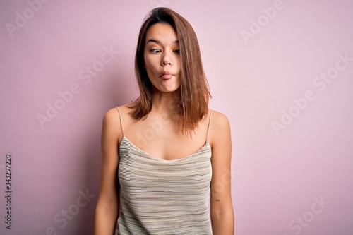 Young beautiful brunette girl wearing casual striped t-shirt over isolated pink background making fish face with lips, crazy and comical gesture. Funny expression.