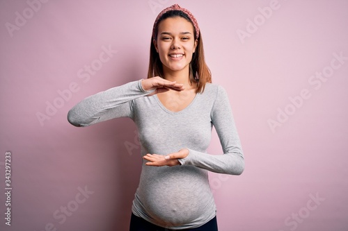 Young beautiful teenager girl pregnant expecting baby over isolated pink background gesturing with hands showing big and large size sign, measure symbol. Smiling looking at the camera. Measuring