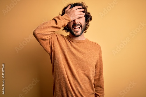 Young handsome man with beard wearing casual sweater standing over yellow background smiling and laughing with hand on face covering eyes for surprise. Blind concept. © Krakenimages.com