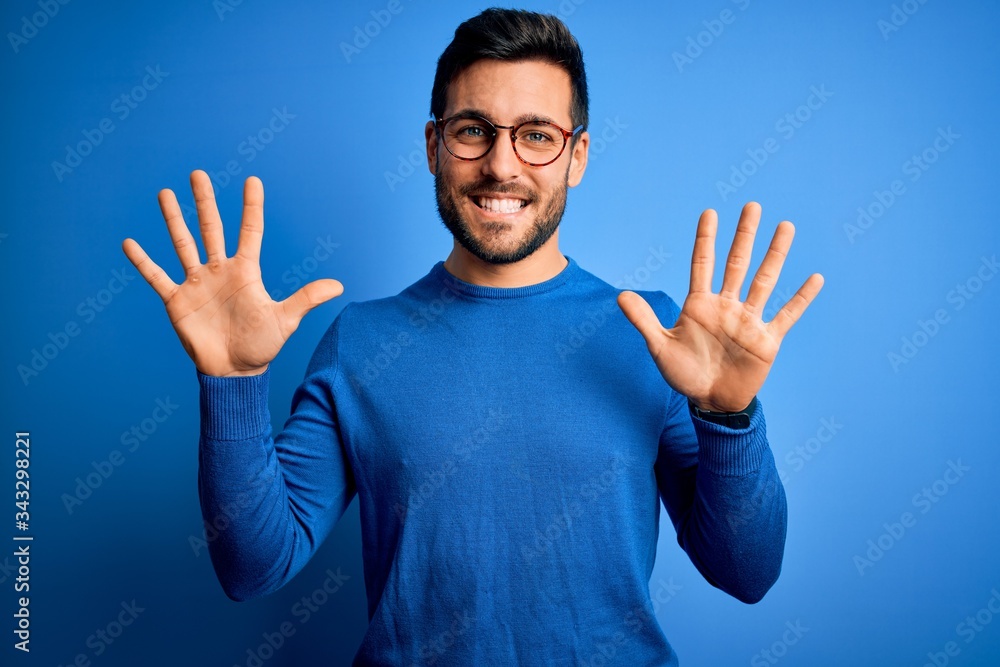 Young handsome man with beard wearing casual sweater and glasses over blue background showing and pointing up with fingers number ten while smiling confident and happy. Young handsome man with beard wearing casual sweater and glasses over blue background showing and pointing up with fingers number ten while smiling confident and happy.
